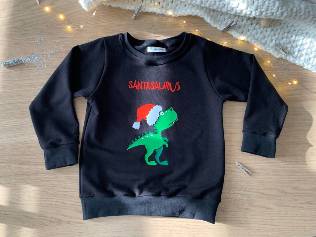Pull de Noël enfant cousu en sweat noir avec motif dinosaure, posé près d’une guirlande